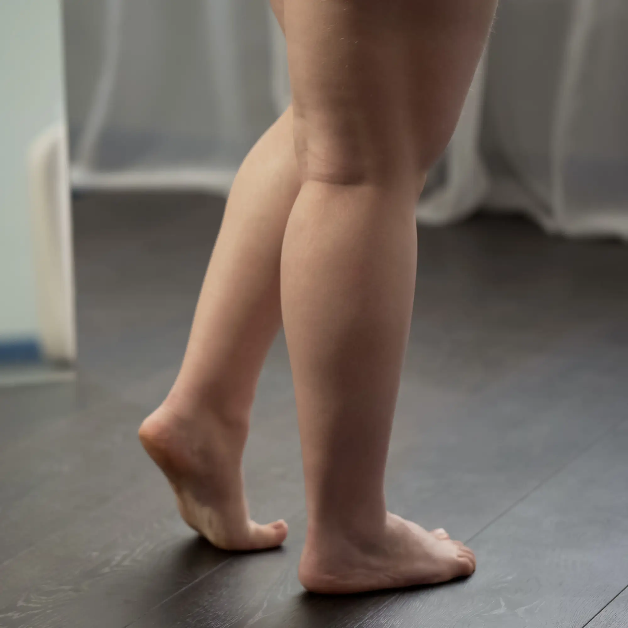 Jambes d'une personne debout sur la pointe des pieds sur un sol en bois foncé, présentant un gonflement suggérant un besoin de drainage lymphatique, avec un rideau flou en arrière-plan.